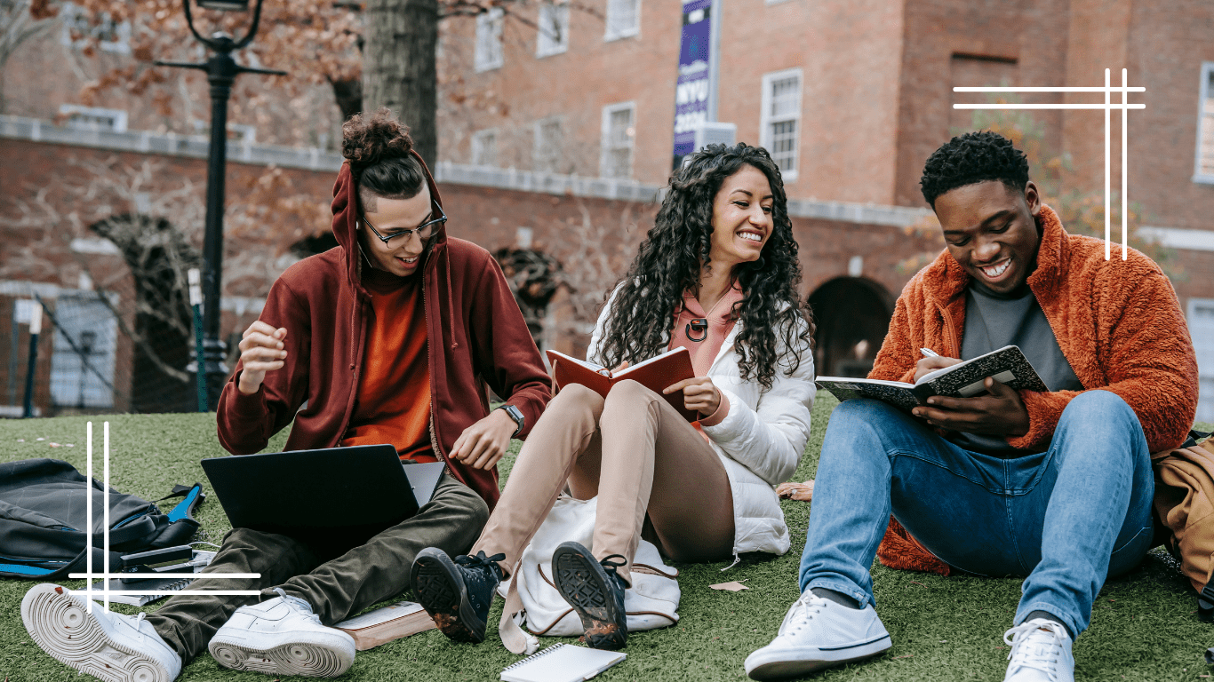 Three college students sit together on grass, smiling and studying. One student works on a laptop wearing a burgundy hoodie, while the others read from books.